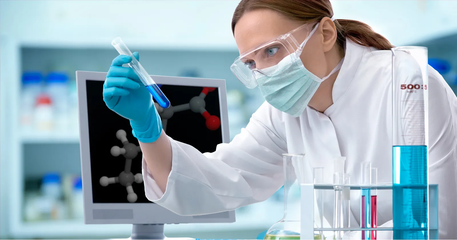 A laboratory researcher in safety glasses and a mask examines a test tube.