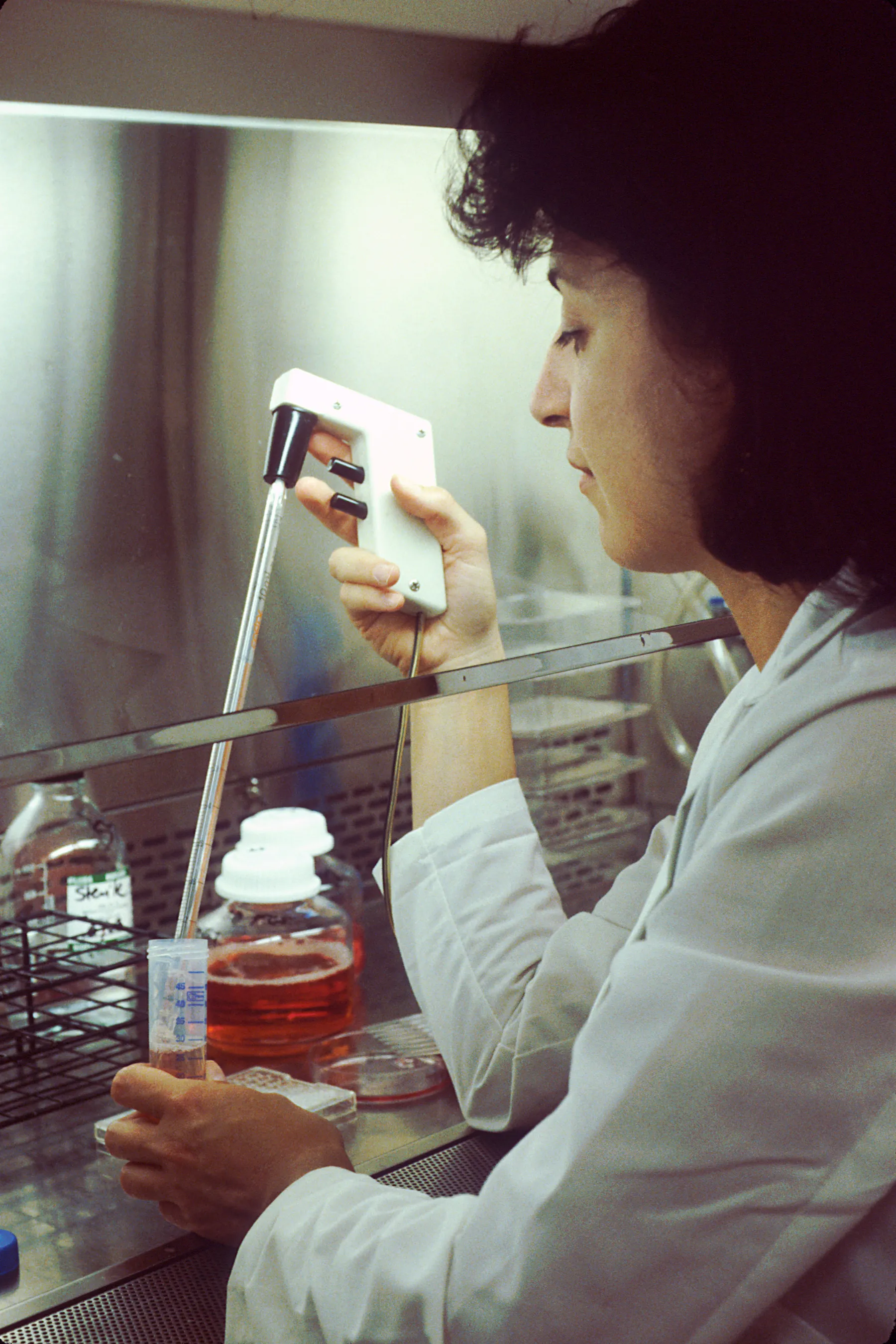A researcher in a lab coat pipetting culture medium inside a biosafety cabinet.